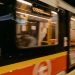 A blurred metro train speeds through an underground station with passengers on board.